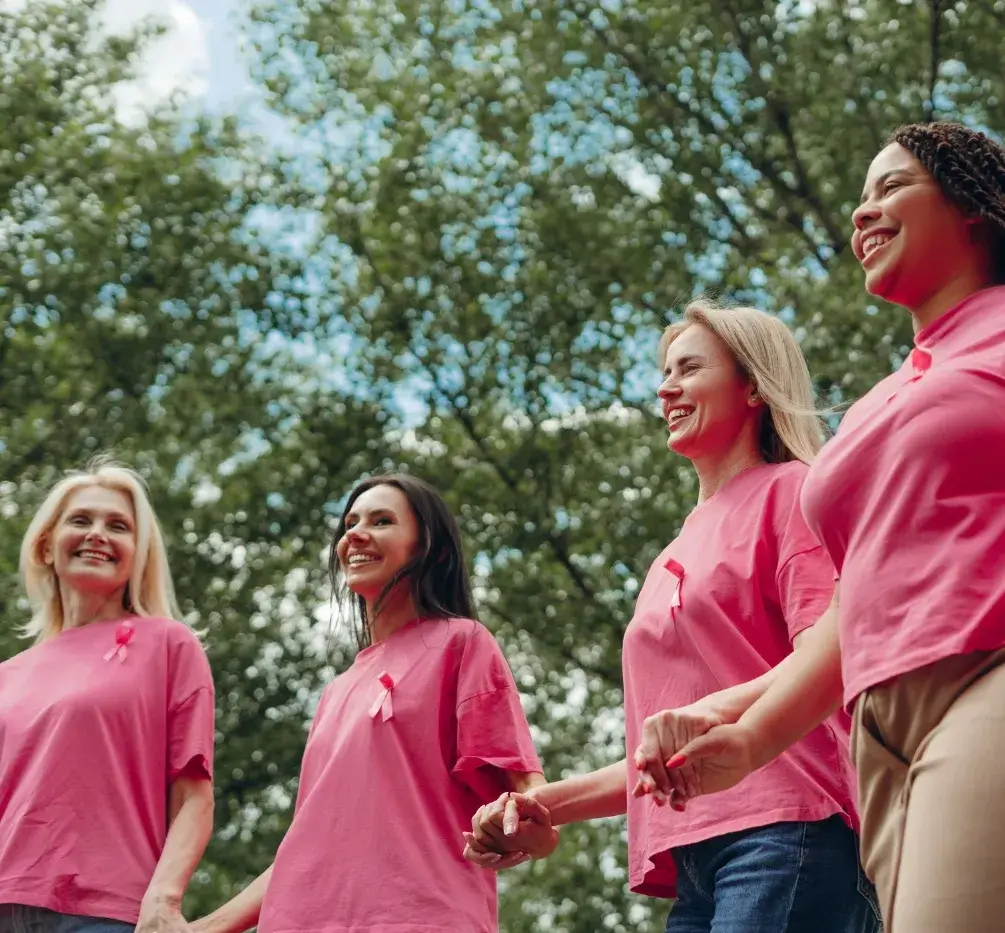 Women wearing pink shirts with breast cancer ribbons, holding hands outdoors in support of Breast Cancer Awareness Month